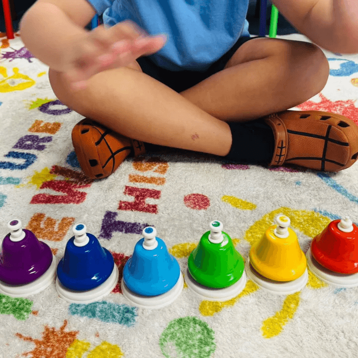 Physical Education and Sports Lessons child sitting in front of a row of colorful handbells.
A close-up view of a child sitting cross-legged on a patterned rug, with a row of seven vibrant, colorful handbells lined up in front: purple, dark blue, light blue, green, yellow, and red. Their hands are in motion—blurred—suggesting interaction with the bells.